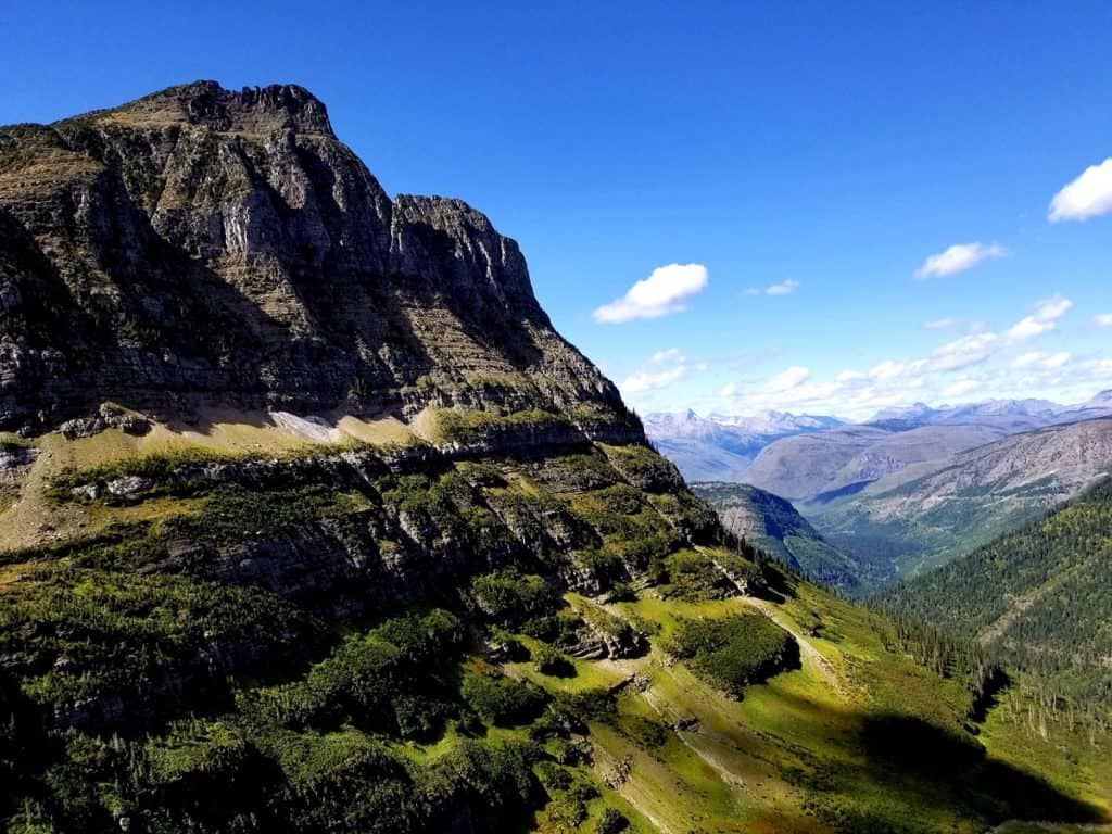 View from Going to the Sun Road in Glacier National Park showing mountain to the left and a large valley with green trees and more mountains in the distance, blue skies with some low clouds