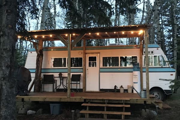 The Marooned Motorhome in West Bragg Creek view of the side of the old motorhome with a deck with cover attached and lights on