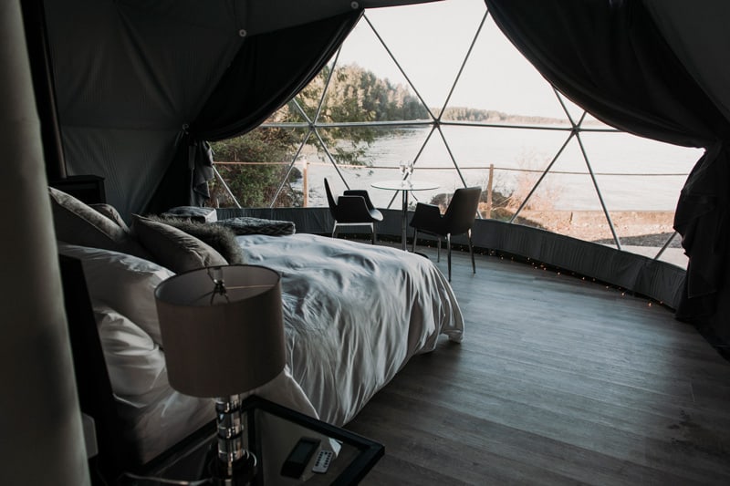 Wild Pods pod glamping in Tofino view from inside with bed, table and chairs, lamp and window view of the ocean
